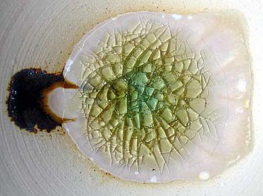 Abb. 5| Gelbes Glas, entstanden durch Sauerstoffeinwirkung bei 1300°C auf eisengrünes Glas. Ein Rest des grünen Glases erhielt sich in der Mitte, der Stelle mit der größten Schichtdicke. - Rings herum Kristallisation und Auswandern des Eisen(III) in den Tonteller. - Links im Bild: Eisenoxidgemisch, entstanden aus einem willentlich zugesetzten Häufchen von Eisen(II)-sulfat. Der Craquelé-Effekt entstand beim Abkühlen. (Versuch und Foto: P. Kurzmann) Abb. 5| Gelbes Glas, entstanden durch Sauerstoffeinwirkung bei 1300°C auf eisengrünes Glas. Ein Rest des grünen Glases erhielt sich in der Mitte, der Stelle mit der größten Schichtdicke. - Rings herum Kristallisation und Auswandern des Eisen(III) in den Tonteller. - Links im Bild: Eisenoxidgemisch, entstanden aus einem willentlich zugesetzten Häufchen von Eisen(II)-sulfat. Der Craquelé-Effekt entstand beim Abkühlen. (Versuch und Foto: P. Kurzmann)