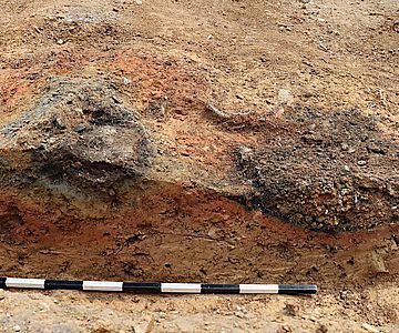 Links ist der Boden des ehemaligen Ofens (grau) erkennbar. Rechts daneben befindet sich eine Grube, die dem Ofen vorgelagert ist. In diese floss Schlacke aus dem Ofen ab