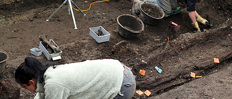 Winterthur. Ausgrabung heute. Freilegung eines Holzkanals (Bildmitte) am Oberwinterthurer Kastellweg im Jahr 2007 durch Mitarbeitende der Kantonsarchäologie. Parallel zum Holzkanal sind noch die Überreste eines Zauns zu sehen. Die blauen Schildchen markieren Schichten, die roten bezeichnen die einzelnen Hölzer. In den grauen Behältern liegt Fundmaterial. (Foto: Kantonsarchäologie Zürich)