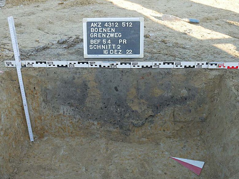 Im Profil ist das Brandgrubengrab anhand einer grau-schwarze Verfüllung in einer flachen Grube gut zu erkennen